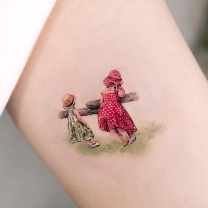 Tatuajes bonitos, pequeños y femeninos; tatuaje de retrato de niñas en el campo con vestidos verde y rojo, en el brazo