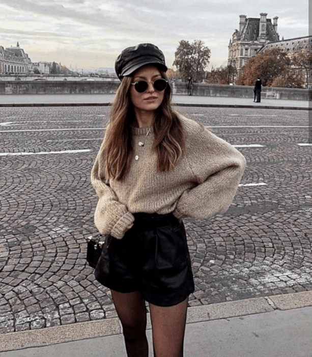 chica de cabello claro largo usando lentes de sol, gorro negro de cuero, suéter beige holgado, shorts de cuero negro, medias negras