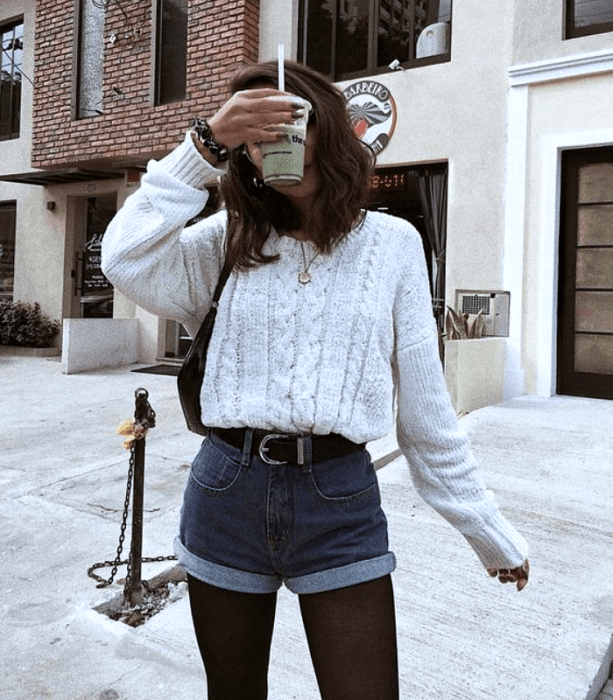 chica de cabello castaño usando un suéter blanco, shorts de mezclilla, medias negras, cinturón negro y bolso de mano negro