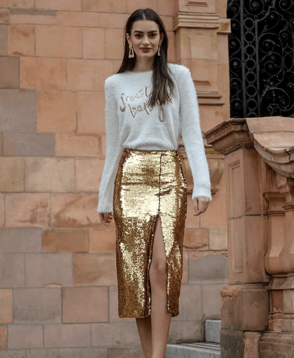 chica de cabello castaño usando una blusa blanca con dorado, falda dorada metálica con apertura en la pierna