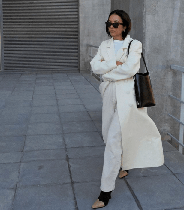 chica de cabello castaño usando lentes de sol, camiseta blanca, abrigo largo blanco, pantalones blancos de mezclilla, botines negros con punta beige y bolso negro de mano