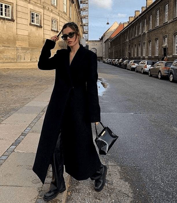 chica de cabello castaño usando lentes de sol, camiseta negra, abrigo largo negro, pantalones de cuero negros, bolso de mano negro y botas de suela gruesa negras