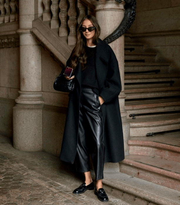 chica de cabello castaño largo usando lentes de sol negros, suéter negro, camiseta blanca, pantalones de cuero, abrigo largo negro, zapatos de piso negros y bolso de mano negro