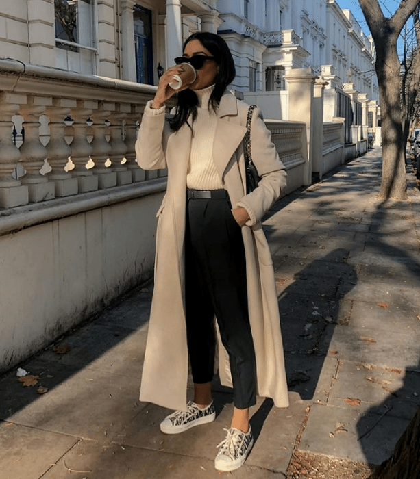 chica de cabello castaño usando lentes de sol, suéter blanco de cuello alto, abrigo largo beige, pantalones de vestir negros, tenis blancos y bolso negro de cuero