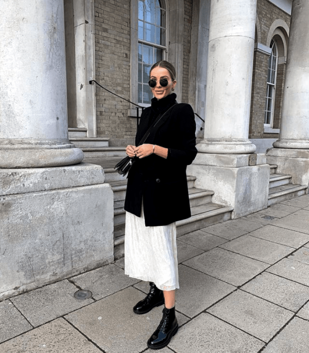 chica rubia usando lentes de sol, abrigo negro, suéter negro de cuello alto, falda blanca larga, botines de cuero de suela gruesa y bolso de mano negro