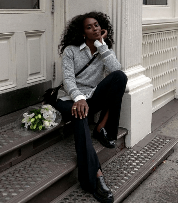 chica morena de cabello chino con camisa blanca de vestir, suéter gris, pantalones de vestir negros, zapatos negros de cuero y bolso negro de cuero