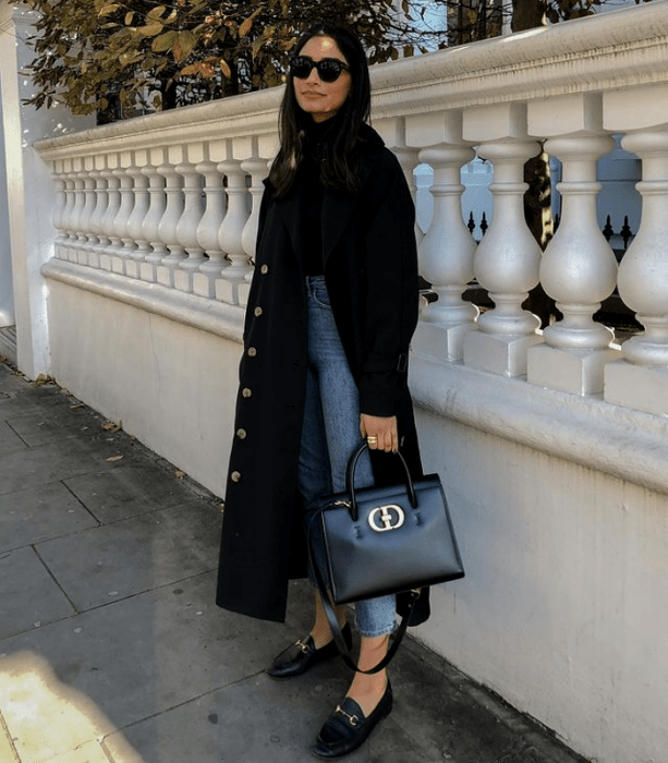 chica de cabello castaño usando lentes de sol, abrigo largo negro, suéter negro de cuello alto, jeans a la cintura rectos, zapatos tipo loafers negros de cuero y bolso de mano negro