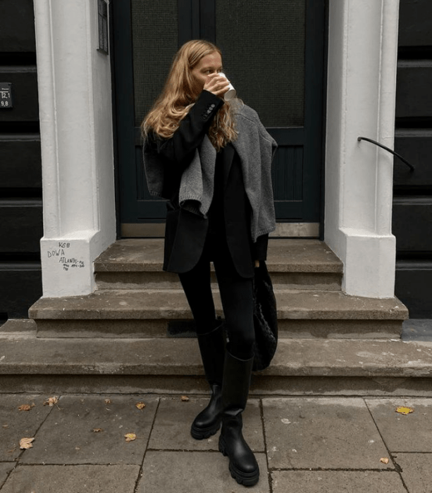 chica rubia usando un saco negro, suéter gris, camiseta negra, leggings negros, botas largas de suela gruesa, bolso de mano negro