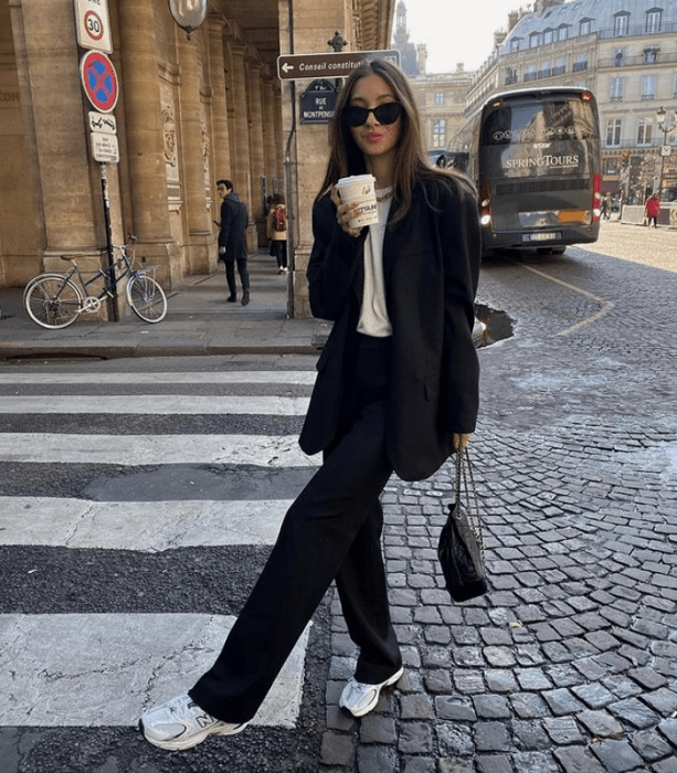 chica de cabello castaño claro usando lentes de sol, camiseta blanca, saco oversized negro, pantalones de vestir negros, tenis blancos y bolso de mano negro