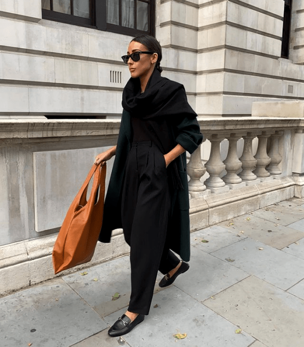 chica morena usando lentes de sol, bufanda negra larga, camiseta negra, abrigo largo verde, pantalones de vestir negros, zapatos negros de piso, bolso de mano naranja