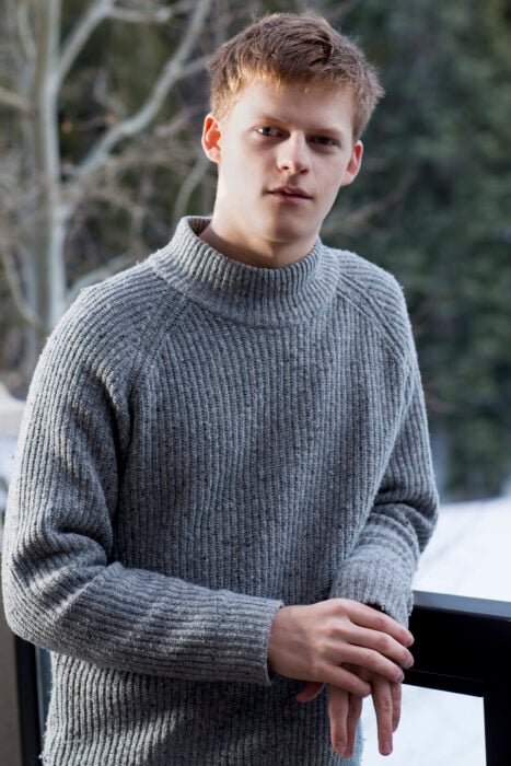 Lucas Hedges llevando suéter en color gris, recargado en una banderilla de madera