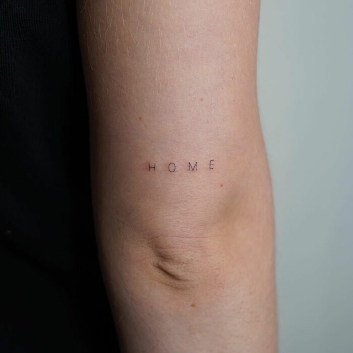 Tatuaje de palabra "home" arriba del codo 