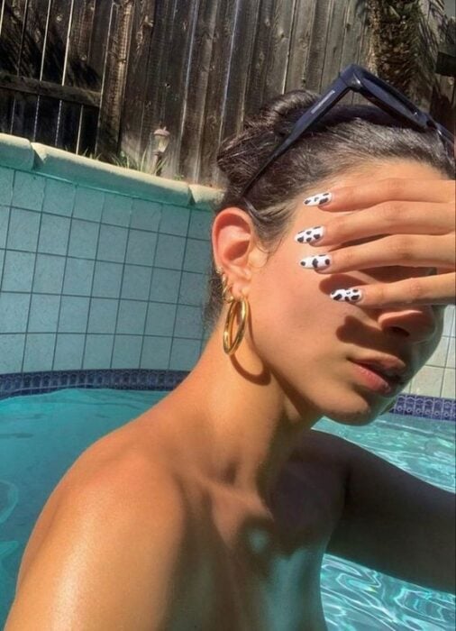 Chica morena clara en la piscina tapándose la cara del sol con su mano y uñas con diseño de vaca