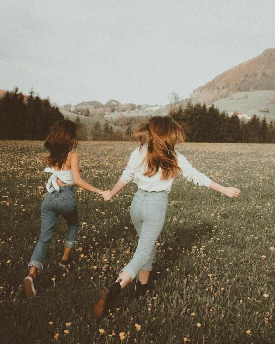 Chica de cabello castaño y chica rubia corriendo por un campo lleno de flores y una  montaña al fondo