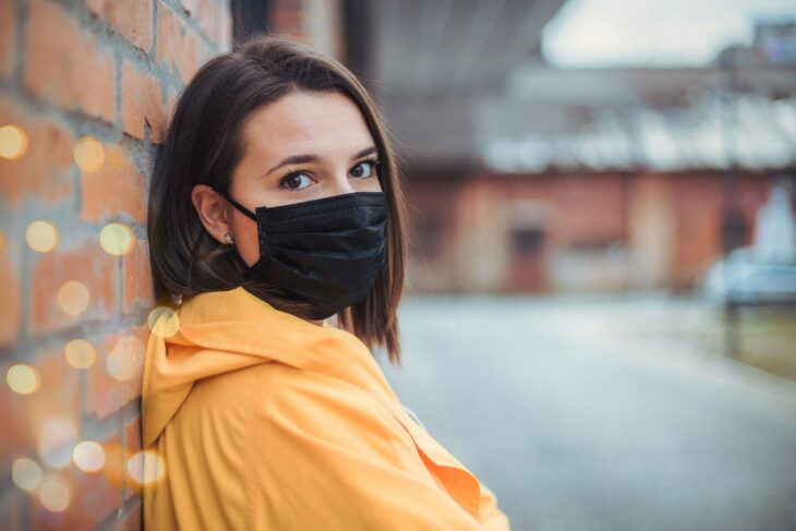 Chica de cabello corto recargada en una pared usando una sudadera amarilla y un cubrebocas negro