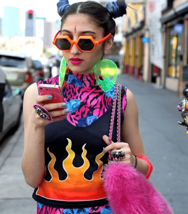 chica de cabello castaño usando lentes de sol naranjas, aretes verdes de alien, top gráfico multicolor, accesorios de plástico, anillos, lentes, aretes