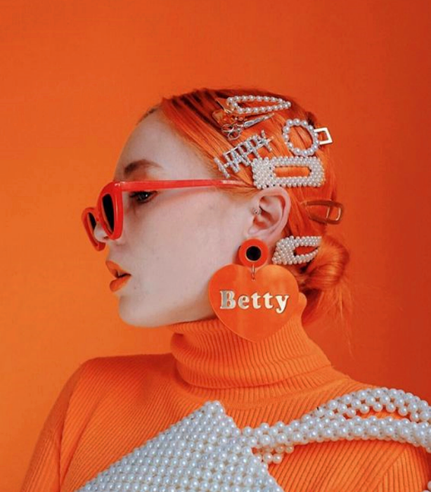 chica de cabello naranja neón, lentes de sol de plástico color naranja, suéter naranja de cuello alto, bolso de cuentas blancas, broches para el cabello de color blanco y naranja de plástico