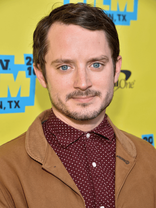 Elijah Wood usando una camisa guinda y saco camel