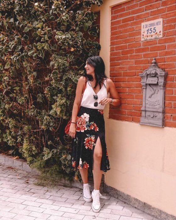 Chica usando una blusa blanca de tirantes, con falda negra de estampado de rosas y tenis blancos