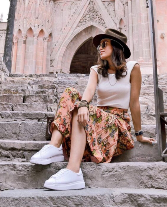 Chica usando blusa color blanco con falda de color naranja con detalles de color verde, tenis blancos y sombrero color negro