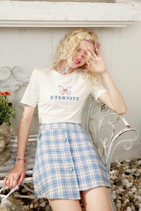 Chica usando una playera blanca con estampado y falda a cuadros de color azul cielo y blanco