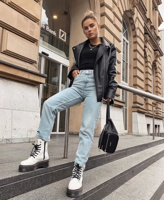 Chica usando jeans, chamarra de cuero, blusa negra y botas Dr. Martens de color blanco con cintas negras 