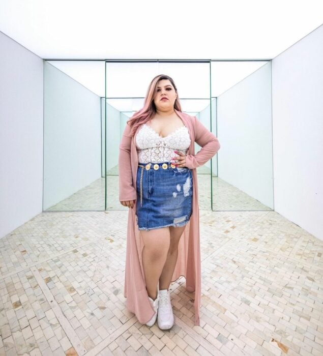 Priscila Arias la Fatshionista usando una falda de jean, top blanco, botas blancas y un ensamble rosa mientras modela 