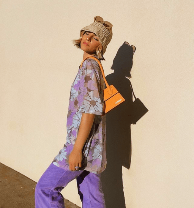 chica de cabello corto rubio usando un bucket hat de flores, blusa oversized lila con flores rosas y pantalones morados, bolso naranja pequeño