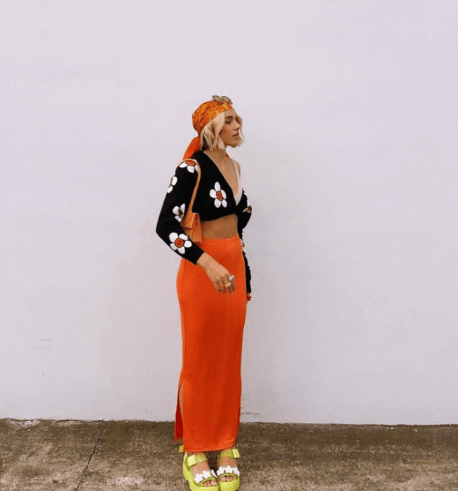 chica rubia usando lentes de sol, blusa negra con flores, falda midi naranja abierta, sandalias amarillas de tacón y mascada en la cabeza