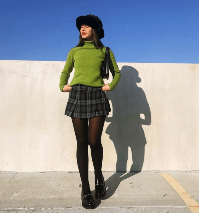 chica de cabello oscuro usando un gorro negro bucket hat, suéter verde, minifalda azul de cuadros, medias negras, botines negros