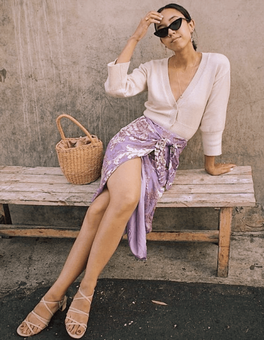 chica de cabello oscuro usando lentes de sol, blusa blanca de manga larga, falda larga lila abierta con flores y sandalias nude de tacón