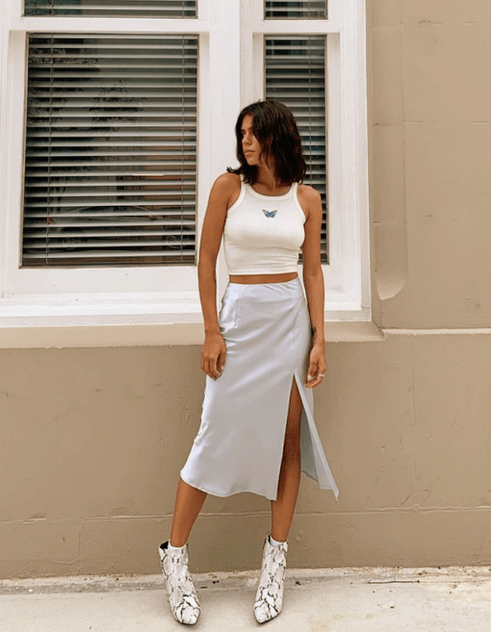 chica morena con crop top blanco de tirantes, falda larga abierta de satén celeste y botines plateados de tacón