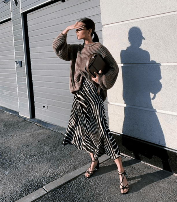 chica de cabello oscuro largo usando lentes de sol, suéter café tejido, falda midi de satén de cebra, sandalias de tacón y bolso de mano cafés