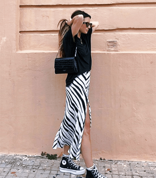 chica de cabello claro largo usando lentes de sol, camiseta oversized negra, falda midi de cebra, tenis converse negros y bolso negro pequeño