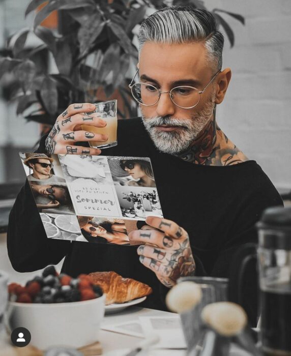 Hombre con tatuajes tomando un desayuno mientras lee una revista 