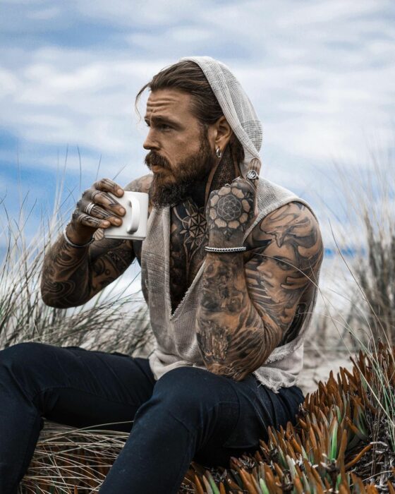 Hombre tatuado tomando una taza de café mientras está admirando el paisaje 