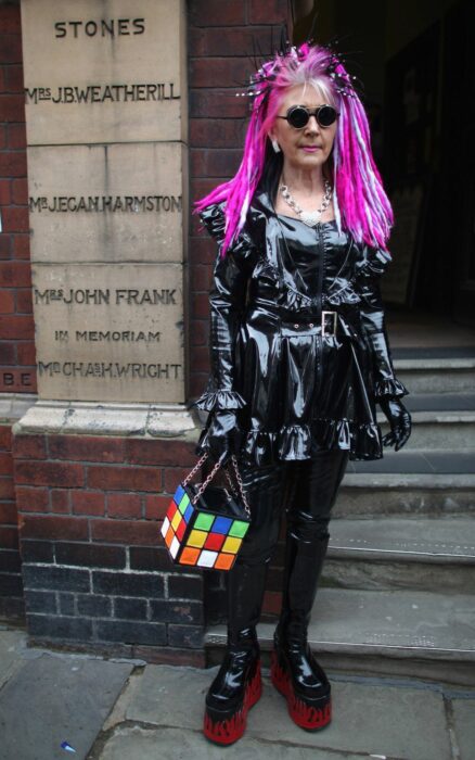 Mujer de la tercera edad vestida de color negro con el cabello rosa y botas de color rojo 