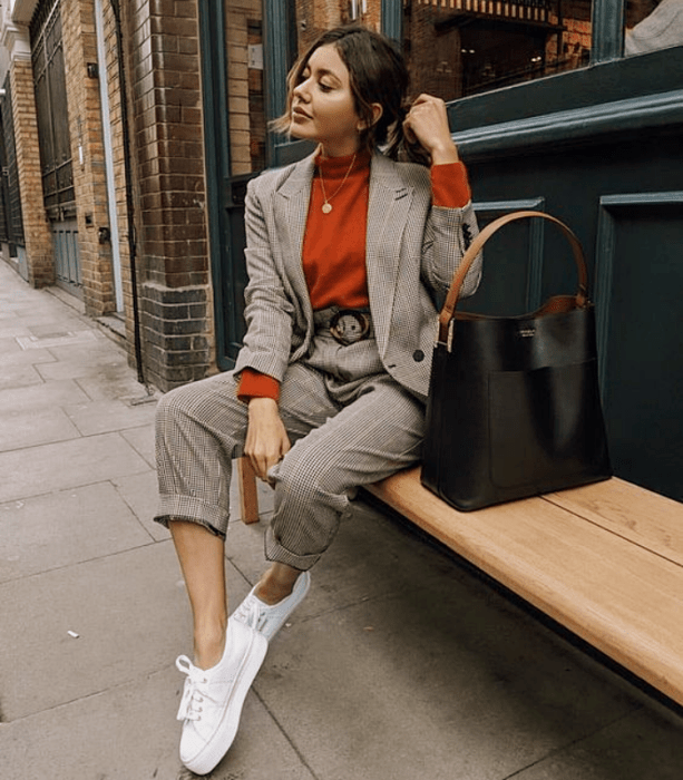 chica morena con cabello castaño usando un top naranja de manga larga, traje gris de cuadros, tenis blancos, bolso negro de cuero