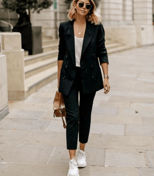 chica de cabello rubio con lentes de sol, camisa blanca, traje negro, tenis blancos, bolso café de piel