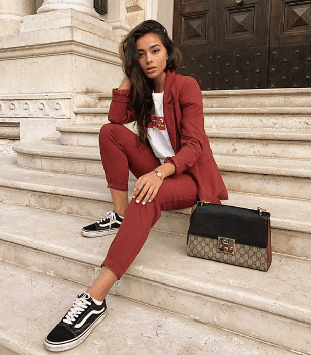 chica de cabello oscuro largo, playera blanca con logo rojo, traje rojo quemado, tenis vans negros con blanco, bolso de mano café con negro de cuero