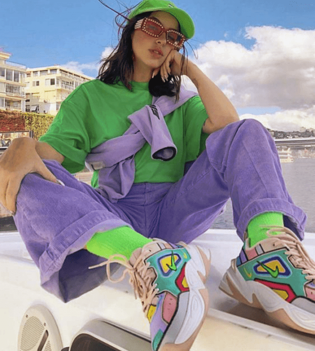 chica de cabello largo castaño usando una gorra de beisbol verde neón, camisa oversized neón, pantalón morado de pana, calcetas neón, cangurera lila, tenis blancos con azul, rosa, rojo y amarillo