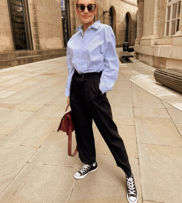 chica rubia con lentes de sol negros, camisa azul celeste de vestir, pantalones negros de vestir, tenis converse negros de botines, bolso café de piel