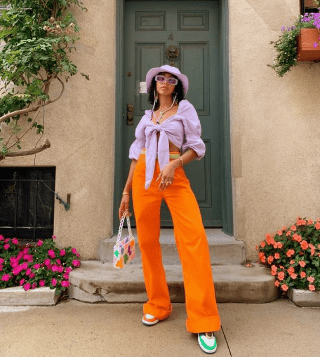 chica morena con cabello largo usando un bucket hat lila, blusa lila de mangas abombadas, pantalón naranja brillante, tenis blancos con naranja y verde