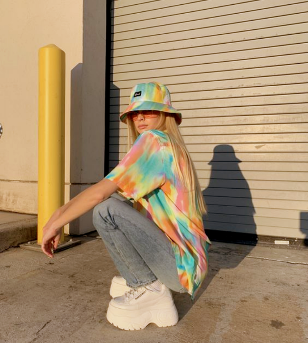 chica rubia con bucket hat de colores de arcoíris, tenis blancos de plataforma, jeans azules holgados y camiseta holgada de tie dye