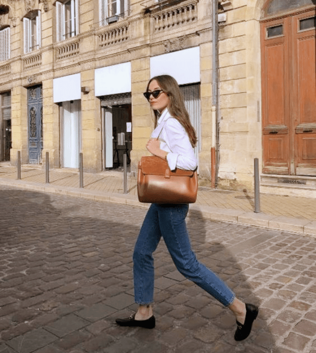 chica de cabello claro con lentes de sol, camisa blanca de vestir, jeans ajustados, flats negros y bolso café de piel