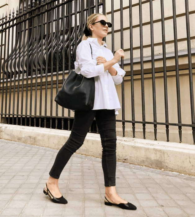 chica rubia con lentes de sol, camisa blanca oversized, jeans negros ajustados, flats negros y bolso negro de cuero