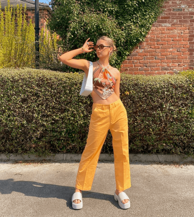 chica de cabello claro usando lentes de sol, top naranja con flores amarillas, pantalones amarillos rectos, sandalias slides blancas, bolso pequeño blanco