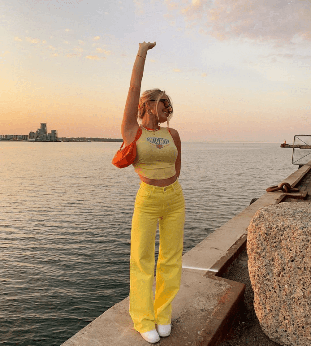 chica de cabello rubio usando un crop top amarillo con cuello halter, pantalones amarillos brillantes, tenis blancos, bolso naranja pequeño