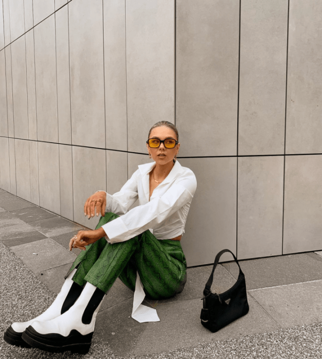 chica de cabello claro con lentes de sol naranjas, blusa blanca de manga larga, pantalones verdes de cuero, botines blancos con negros y bolsa pequeña negra de cuero