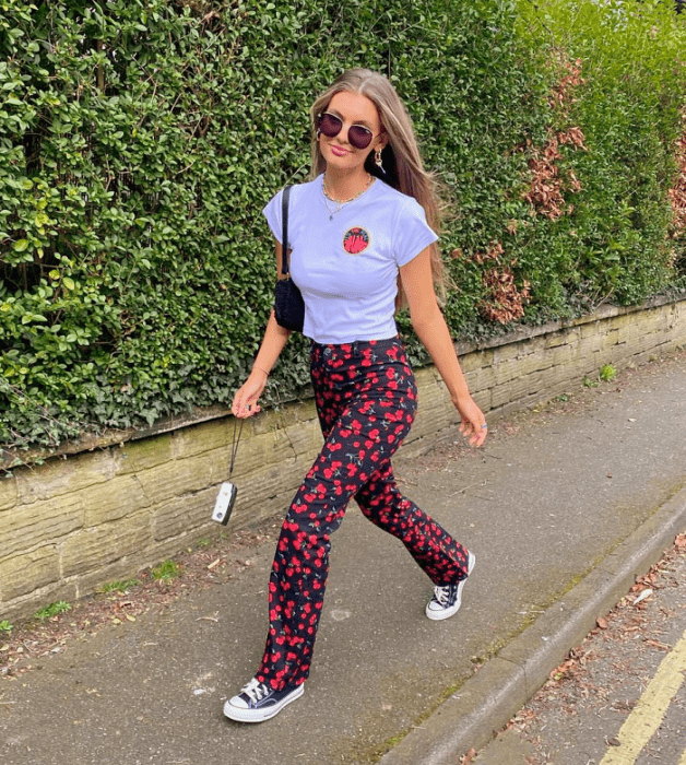 chica de cabello claro usando lentes de sol, camiseta blanca, pantalones negros con flores rojas y cerezas, converse negros con azul